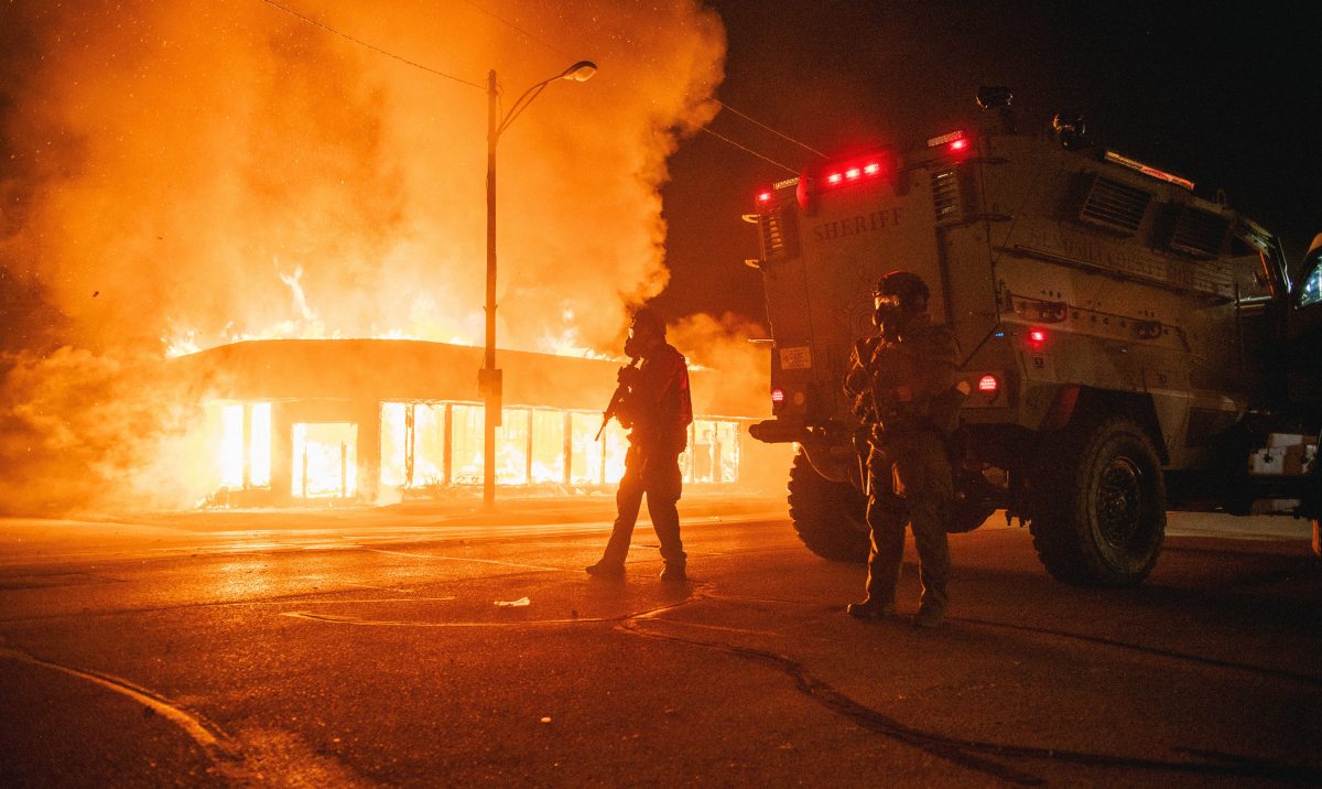 A police armored vehicle patrols an intersection while a building set afire